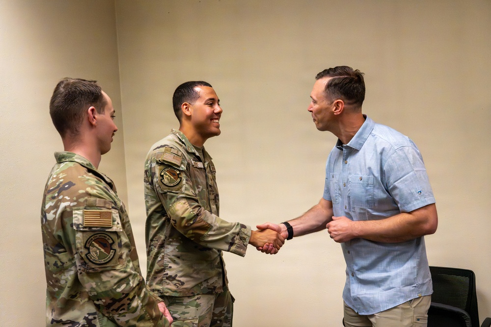 Two military personnel in OCP (Operational Camouflage Pattern) uniforms are meeting with a civilian man in a light blue shirt. The central uniformed individual is shaking hands with the civilian while smiling, suggesting a formal greeting or recognition event. The setting appears to be an indoor office or meeting room with plain beige walls.