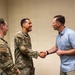 Two military personnel in OCP (Operational Camouflage Pattern) uniforms are meeting with a civilian man in a light blue shirt. The central uniformed individual is shaking hands with the civilian while smiling, suggesting a formal greeting or recognition event. The setting appears to be an indoor office or meeting room with plain beige walls.
