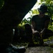 A person is crouching in a dense jungle or tropical forest environment, handling yellow straps or rope on the ground. They are wearing olive/military-style clothing including a bucket hat, t-shirt, and tactical gloves. The scene is dramatically lit with dappled light filtering through the forest canopy.