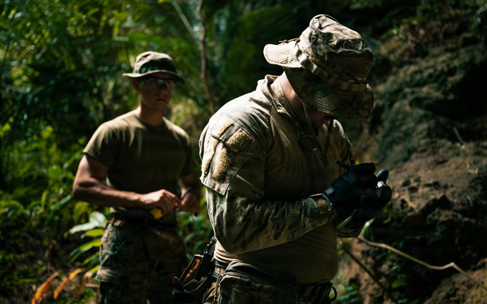 Two military personnel are operating in a dense jungle or forested environment. The foreground figure is wearing full camouflage combat gear including a boonie hat and tactical gloves, and appears to be examining or operating a small handheld device. The background figure is in a green t-shirt and camouflage trousers with sunglasses, holding what appears to be a small yellow object.