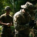Two military personnel are operating in a dense jungle or forested environment. The foreground figure is wearing full camouflage combat gear including a boonie hat and tactical gloves, and appears to be examining or operating a small handheld device. The background figure is in a green t-shirt and camouflage trousers with sunglasses, holding what appears to be a small yellow object.