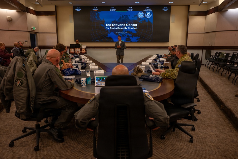 A briefing is being conducted at the Ted Stevens Center for Arctic Security Studies, as shown on the large display screen at the front of the room. Military personnel and civilians are seated around a large oval conference table, listening to a presenter standing at the front. The setting appears to be a formal military conference room with multiple time zone clocks displayed above the main screen.