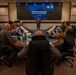A briefing is being conducted at the Ted Stevens Center for Arctic Security Studies, as shown on the large display screen at the front of the room. Military personnel and civilians are seated around a large oval conference table, listening to a presenter standing at the front. The setting appears to be a formal military conference room with multiple time zone clocks displayed above the main screen.