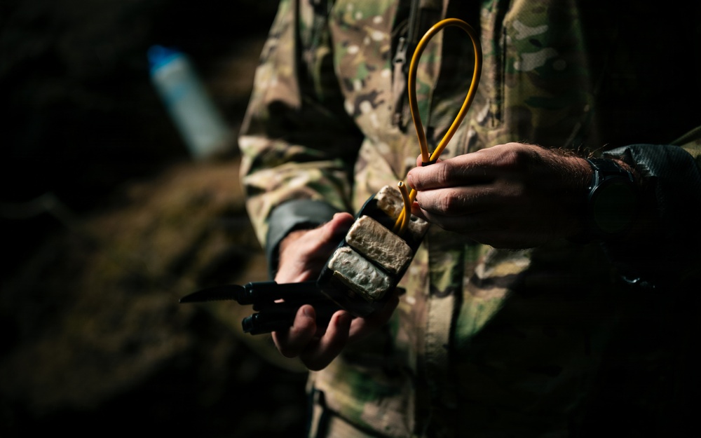 A person wearing military camouflage clothing is handling what appears to be a demolition training device or explosive charge with detonating cord (det cord) attached. The individual is also holding a tool, likely pliers or a multi-tool, in the other hand. This appears consistent with military explosive ordnance or combat engineering training.