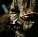A person wearing military camouflage clothing is handling what appears to be a demolition training device or explosive charge with detonating cord (det cord) attached. The individual is also holding a tool, likely pliers or a multi-tool, in the other hand. This appears consistent with military explosive ordnance or combat engineering training.