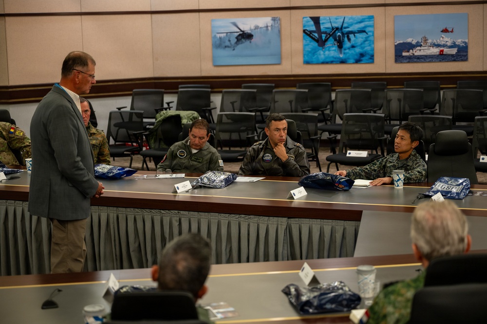 A briefing or meeting is taking place in a military conference room with multinational military personnel seated at a long table. A civilian in a gray suit is standing and addressing the group, which includes personnel in various military uniforms from different nations. Aviation and maritime photographs are displayed on the wall in the background.