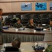 A briefing or meeting is taking place in a military conference room with multinational military personnel seated at a long table. A civilian in a gray suit is standing and addressing the group, which includes personnel in various military uniforms from different nations. Aviation and maritime photographs are displayed on the wall in the background.