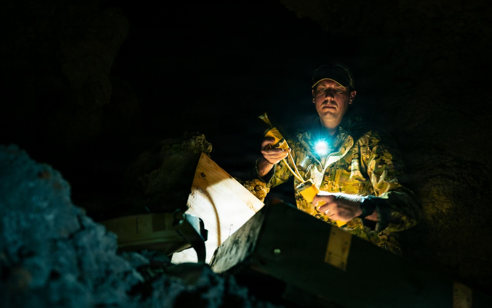 A man wearing camouflage clothing and a dark cap is working in a dark cave or underground environment, illuminated by a bright headlamp or chest-mounted light. He appears to be handling wooden box structures, possibly bat boxes or wildlife monitoring equipment. The dramatic low-key lighting creates strong contrast between the illuminated subject and the surrounding darkness.