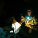A man wearing camouflage clothing and a dark cap is working in a dark cave or underground environment, illuminated by a bright headlamp or chest-mounted light. He appears to be handling wooden box structures, possibly bat boxes or wildlife monitoring equipment. The dramatic low-key lighting creates strong contrast between the illuminated subject and the surrounding darkness.
