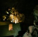 A person in camouflage or military-style clothing is navigating or climbing through a dark cave or underground tunnel environment, illuminated by artificial light. At least one firearm is visible on the cave floor below. A second individual in camouflage is partially visible and out of focus in the foreground right.