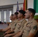 Four U.S. Marine Corps service members in tan service uniforms are seated in a row inside what appears to be a military chapel, attending a formal ceremony. They are facing the same direction, suggesting they are watching a presentation or ceremony in progress. Multiple service ribbons and rank insignia are visible on their uniforms.