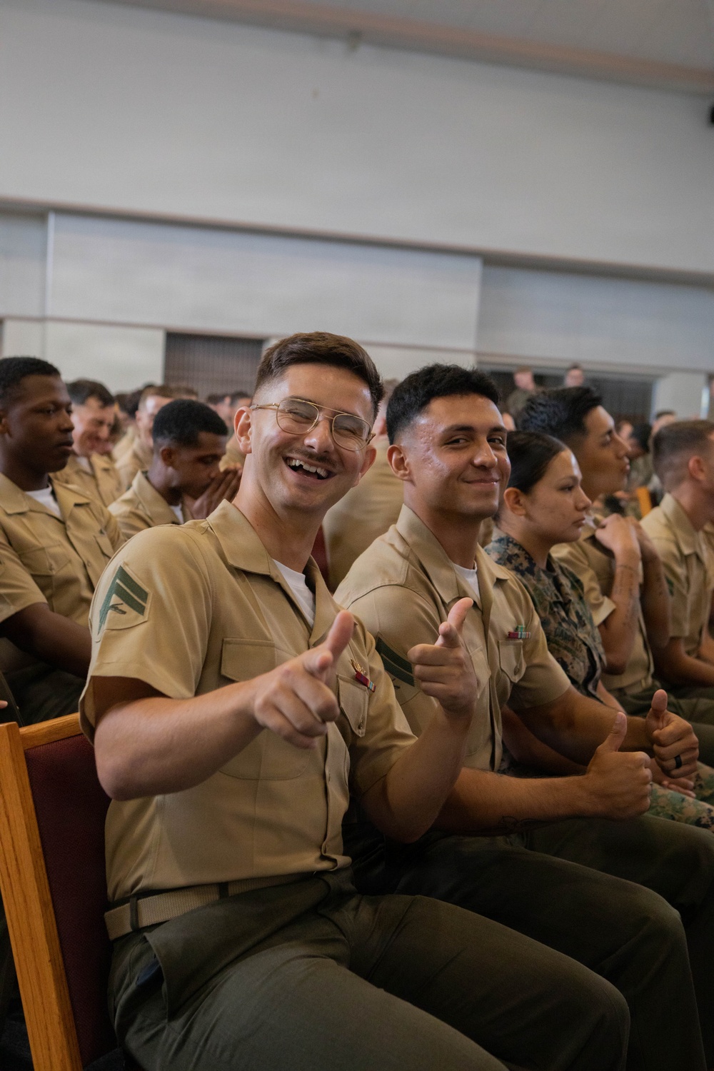 A group of U.S. Marine Corps personnel seated in what appears to be an auditorium or assembly hall during a formal gathering or ceremony. Two Marines in the foreground are smiling and giving thumbs-up/finger-gun gestures toward the camera. The Marines are wearing tan Service 'C' uniforms with olive green trousers.