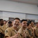 A group of U.S. Marine Corps personnel seated in what appears to be an auditorium or assembly hall during a formal gathering or ceremony. Two Marines in the foreground are smiling and giving thumbs-up/finger-gun gestures toward the camera. The Marines are wearing tan Service 'C' uniforms with olive green trousers.