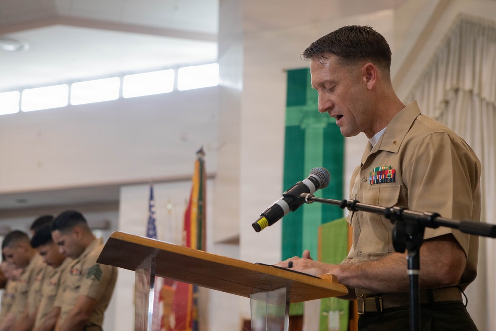 A U.S. Marine Corps officer in service uniform (tan/khaki) is speaking at a podium with a microphone during what appears to be a formal military ceremony or prayer service. Behind him, other Marines in uniform stand with heads bowed, suggesting a moment of prayer or reflection. The setting appears to be a chapel or ceremonial hall with military flags visible in the background.