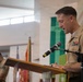 A U.S. Marine Corps officer in service uniform (tan/khaki) is speaking at a podium with a microphone during what appears to be a formal military ceremony or prayer service. Behind him, other Marines in uniform stand with heads bowed, suggesting a moment of prayer or reflection. The setting appears to be a chapel or ceremonial hall with military flags visible in the background.