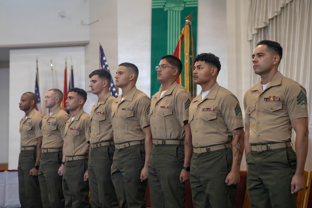 A group of eight United States Marines in tan service uniforms (Charlie or Delta service uniform) stand in a formal line at what appears to be a military ceremony or promotion event. They are positioned in an indoor venue with military flags visible in the background, including the American flag and what appears to be a unit guidon. The Marines are standing at attention, facing to their right.