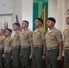 A group of eight United States Marines in tan service uniforms (Charlie or Delta service uniform) stand in a formal line at what appears to be a military ceremony or promotion event. They are positioned in an indoor venue with military flags visible in the background, including the American flag and what appears to be a unit guidon. The Marines are standing at attention, facing to their right.