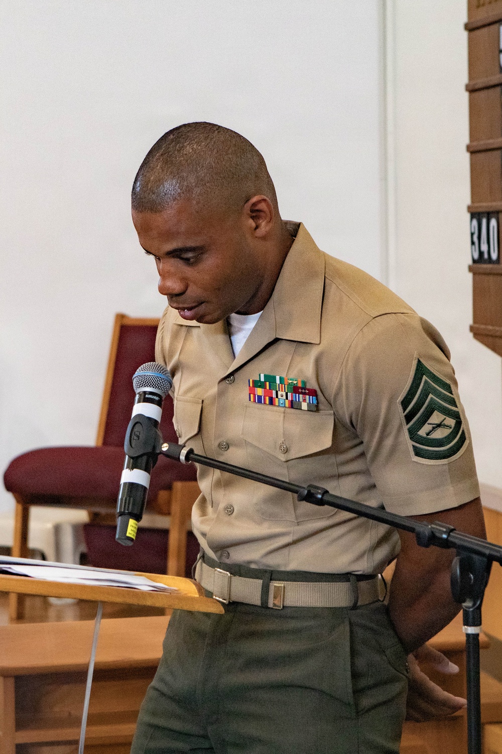 A U.S. Marine in service uniform (tan shirt, olive green trousers) stands at a podium speaking into a Shure microphone. He displays multiple service ribbons on his chest and wears a Staff Sergeant or Gunnery Sergeant rank insignia patch on his left sleeve. The setting appears to be an indoor ceremonial or formal event, possibly in a chapel or meeting hall.