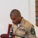 A U.S. Marine in service uniform (tan shirt, olive green trousers) stands at a podium speaking into a Shure microphone. He displays multiple service ribbons on his chest and wears a Staff Sergeant or Gunnery Sergeant rank insignia patch on his left sleeve. The setting appears to be an indoor ceremonial or formal event, possibly in a chapel or meeting hall.