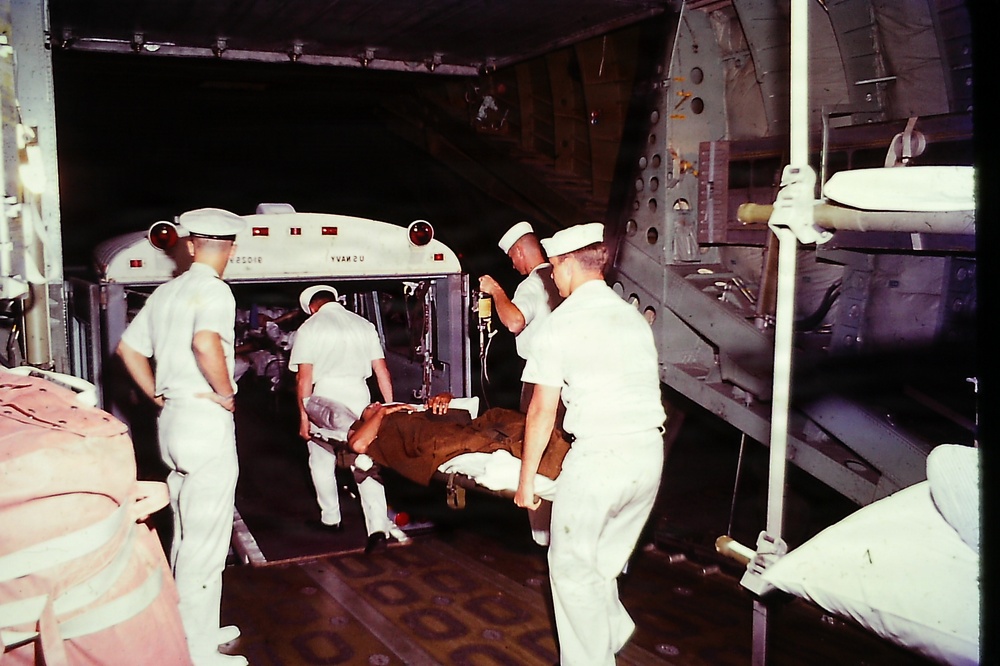 The image shows military medical personnel in white uniforms transferring a patient on a stretcher, likely aboard a large military transport aircraft or ship. An ambulance bus is visible in the background, suggesting this is a medical evacuation operation. An IV bag is visibly attached to the patient, indicating active medical care during transport.