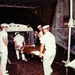 The image shows military medical personnel in white uniforms transferring a patient on a stretcher, likely aboard a large military transport aircraft or ship. An ambulance bus is visible in the background, suggesting this is a medical evacuation operation. An IV bag is visibly attached to the patient, indicating active medical care during transport.