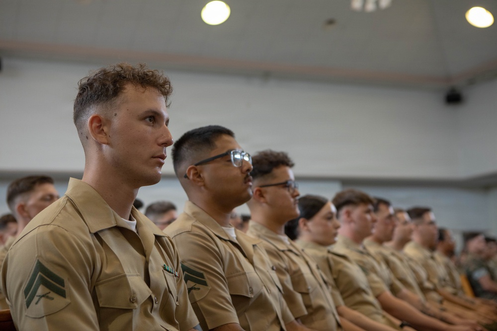 A group of uniformed military personnel, appearing to be U.S. Marines based on their tan/khaki service uniforms and visible rank insignia, are seated in rows in what appears to be an auditorium or briefing room. The individuals are attentively facing forward, suggesting they are attending a briefing, lecture, or formal presentation. The foreground subject displays corporal rank chevrons on their collar.