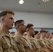 A group of uniformed military personnel, appearing to be U.S. Marines based on their tan/khaki service uniforms and visible rank insignia, are seated in rows in what appears to be an auditorium or briefing room. The individuals are attentively facing forward, suggesting they are attending a briefing, lecture, or formal presentation. The foreground subject displays corporal rank chevrons on their collar.