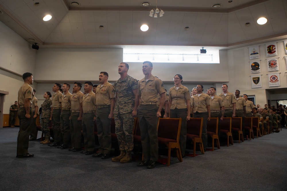 A military ceremony or formation is taking place inside a large indoor hall, likely on a military installation. A group of uniformed service members, appearing to be U.S. Marines in both camouflage and tan/khaki service uniforms, are standing in formation. Several individuals appear to be speaking or reciting something in unison, suggesting a formal ceremony such as a promotion, graduation, or reenlistment.