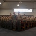 A military ceremony or formation is taking place inside a large indoor hall, likely on a military installation. A group of uniformed service members, appearing to be U.S. Marines in both camouflage and tan/khaki service uniforms, are standing in formation. Several individuals appear to be speaking or reciting something in unison, suggesting a formal ceremony such as a promotion, graduation, or reenlistment.