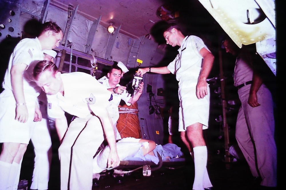 An archival color photograph showing military medical personnel in white uniforms attending to a patient on a stretcher inside what appears to be a military transport aircraft. Several individuals are gathered around the litter, with one person holding what appears to be medical equipment or an IV apparatus. The image has a vintage color cast consistent with mid-20th century film photography, likely from the Vietnam War era.