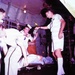 An archival color photograph showing military medical personnel in white uniforms attending to a patient on a stretcher inside what appears to be a military transport aircraft. Several individuals are gathered around the litter, with one person holding what appears to be medical equipment or an IV apparatus. The image has a vintage color cast consistent with mid-20th century film photography, likely from the Vietnam War era.