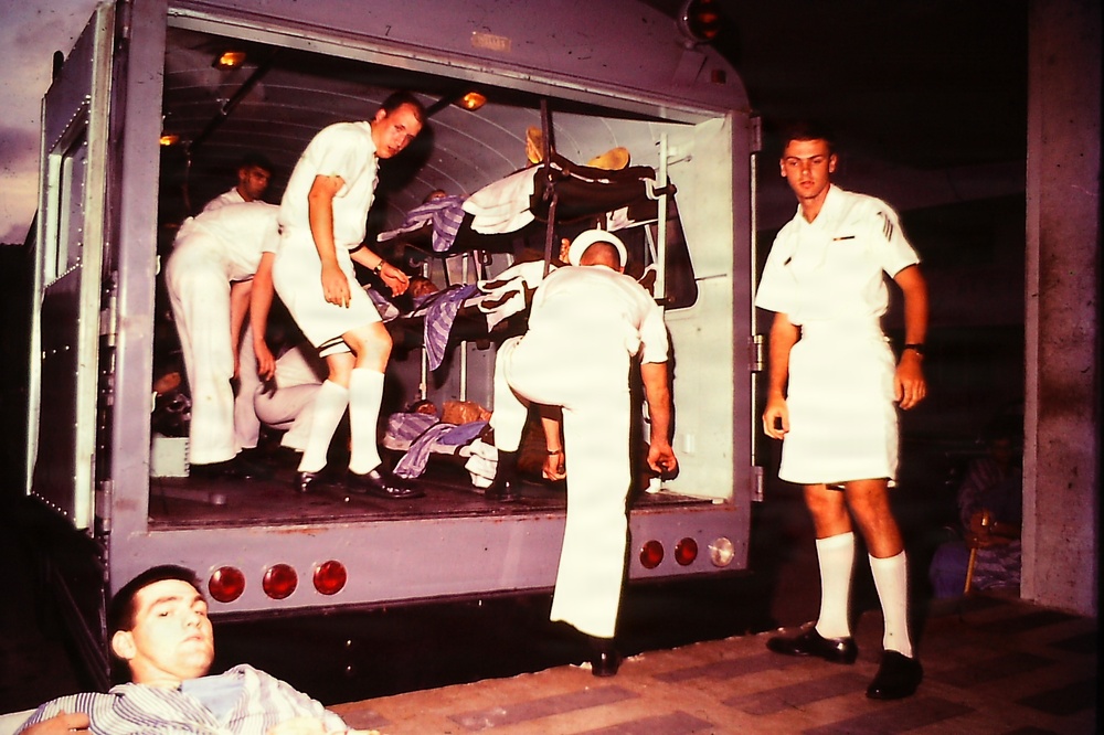 A vintage color photograph showing several individuals in white uniforms, consistent with U.S. Navy or military medical personnel, attending to what appears to be casualties or patients inside and around the rear of a large vehicle, possibly a bus or transport. The scene suggests a medical response or casualty evacuation exercise or real incident. One person in civilian clothes is visible lying in the foreground.