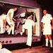 A vintage color photograph showing several individuals in white uniforms, consistent with U.S. Navy or military medical personnel, attending to what appears to be casualties or patients inside and around the rear of a large vehicle, possibly a bus or transport. The scene suggests a medical response or casualty evacuation exercise or real incident. One person in civilian clothes is visible lying in the foreground.