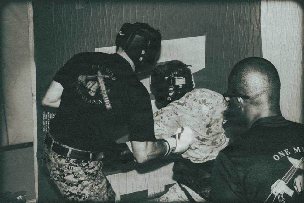 A black-and-white/sepia-toned photograph showing what appears to be a military or law enforcement combatives training session. Two individuals wearing 'One Mind One Weapon' branded black t-shirts are engaged with a third person in camouflage uniform and protective headgear. The setting appears to be an indoor training facility or tent structure.