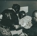 A black-and-white/sepia-toned photograph showing what appears to be a military or law enforcement combatives training session. Two individuals wearing 'One Mind One Weapon' branded black t-shirts are engaged with a third person in camouflage uniform and protective headgear. The setting appears to be an indoor training facility or tent structure.