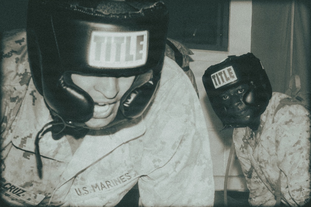Two U.S. Marines wearing TITLE brand boxing headgear are photographed in what appears to be a training or sparring context. Both individuals are dressed in USMC digital camouflage uniforms. The image has a sepia/aged photographic tone with visible text identifying 'U.S. MARINES' and a partial surname 'CRUZ' on one uniform.