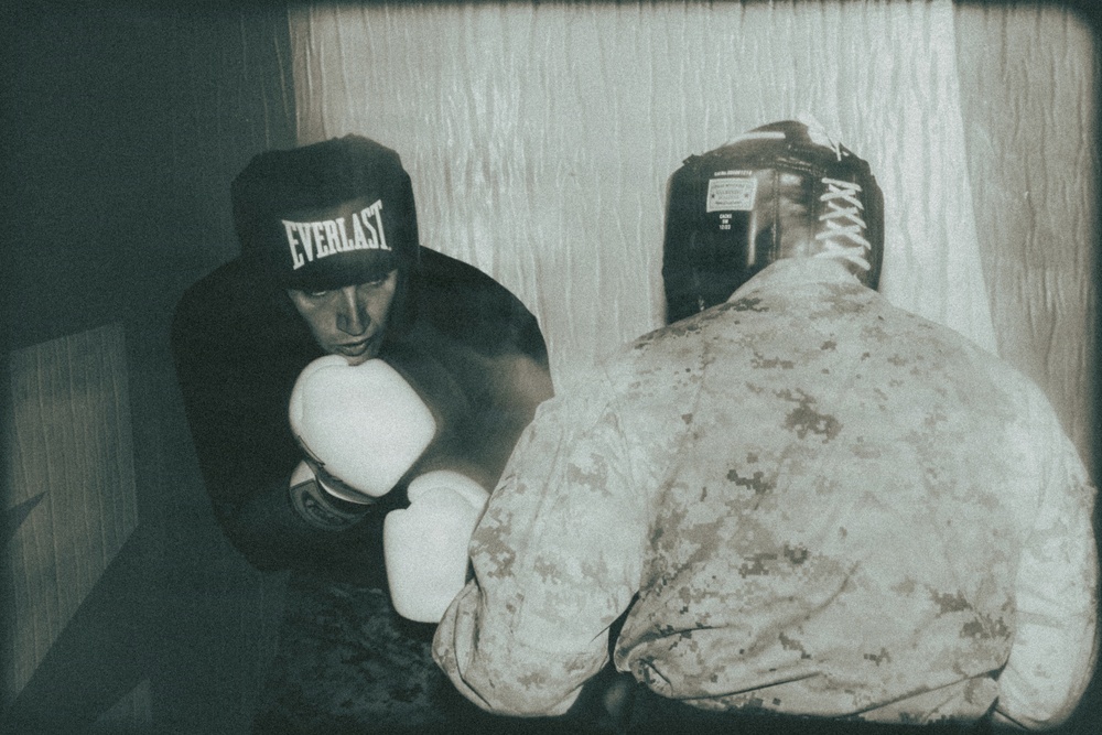 Two individuals are engaged in a boxing sparring session in what appears to be an informal indoor setting. One boxer faces the camera wearing an Everlast branded headgear and white gloves, while the other person, seen from behind, is wearing military camouflage uniform and a black headgear. The image has a grainy, low-light film aesthetic suggesting it was taken with analog or lomography-style photography.