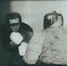 Two individuals are engaged in a boxing sparring session in what appears to be an informal indoor setting. One boxer faces the camera wearing an Everlast branded headgear and white gloves, while the other person, seen from behind, is wearing military camouflage uniform and a black headgear. The image has a grainy, low-light film aesthetic suggesting it was taken with analog or lomography-style photography.