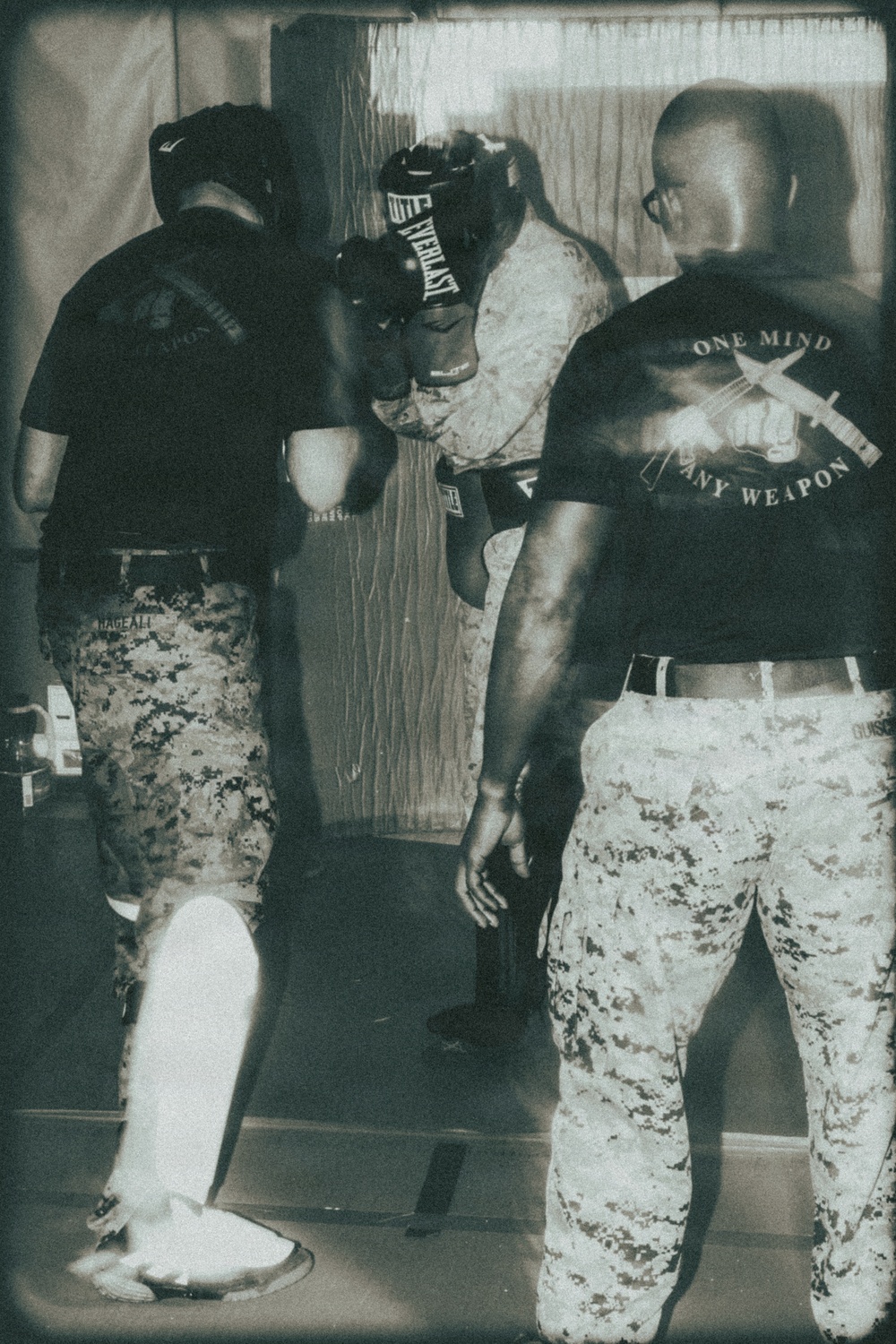 A black-and-white/sepia-toned photograph showing military personnel engaged in a boxing or martial arts training session. One individual wearing an Everlast boxing headgear and gloves appears to be sparring or drilling, while others in USMC digital camouflage (MARPAT) uniforms observe or participate. The setting appears to be an indoor training space, possibly a forward operating base or military facility.