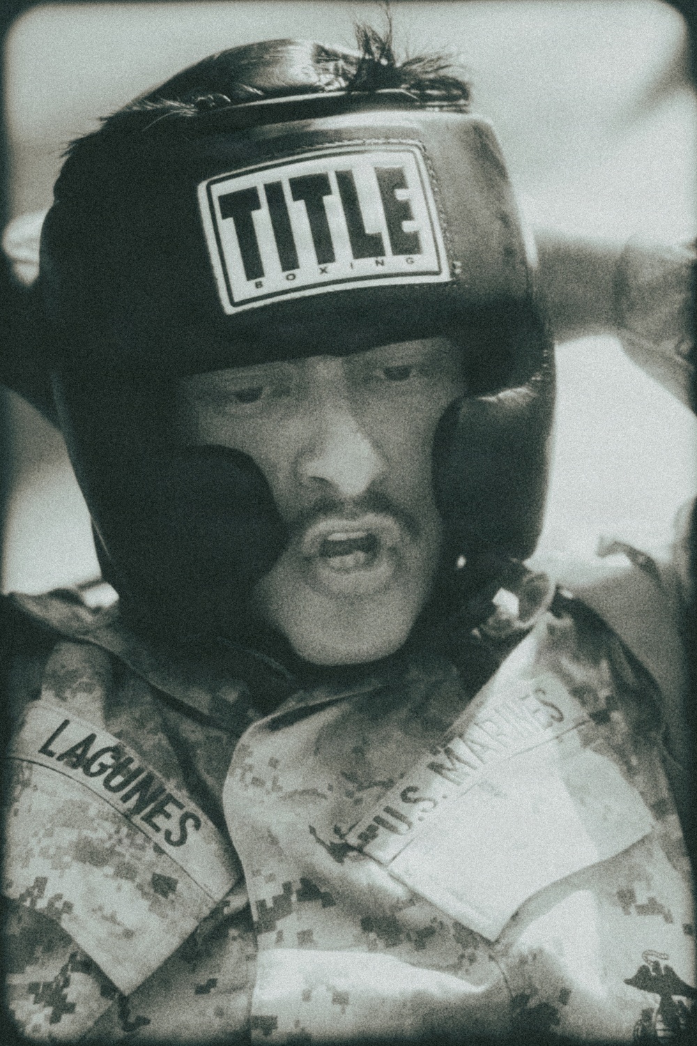 A black-and-white photograph of a U.S. Marine wearing a 'Title Boxing' headgear and digital camouflage (MARPAT) uniform. The individual's name tape reads 'LAGUNES' and the branch tape reads 'U.S. MARINES.' The subject appears to be engaged in a boxing or combatives training session.