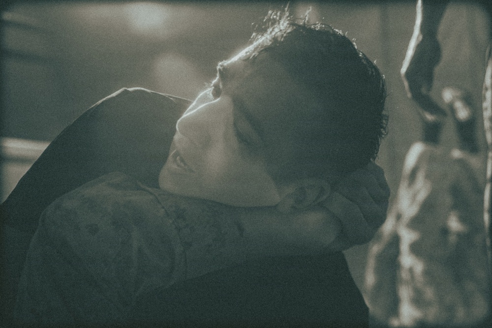 A dramatic, moody photograph of a person with their head tilted back, illuminated by a soft light source from above. The image has a cinematic, dark aesthetic with a desaturated teal-green color grade. The subject appears to be wearing a dark patterned garment and is positioned near a textured surface on the right.