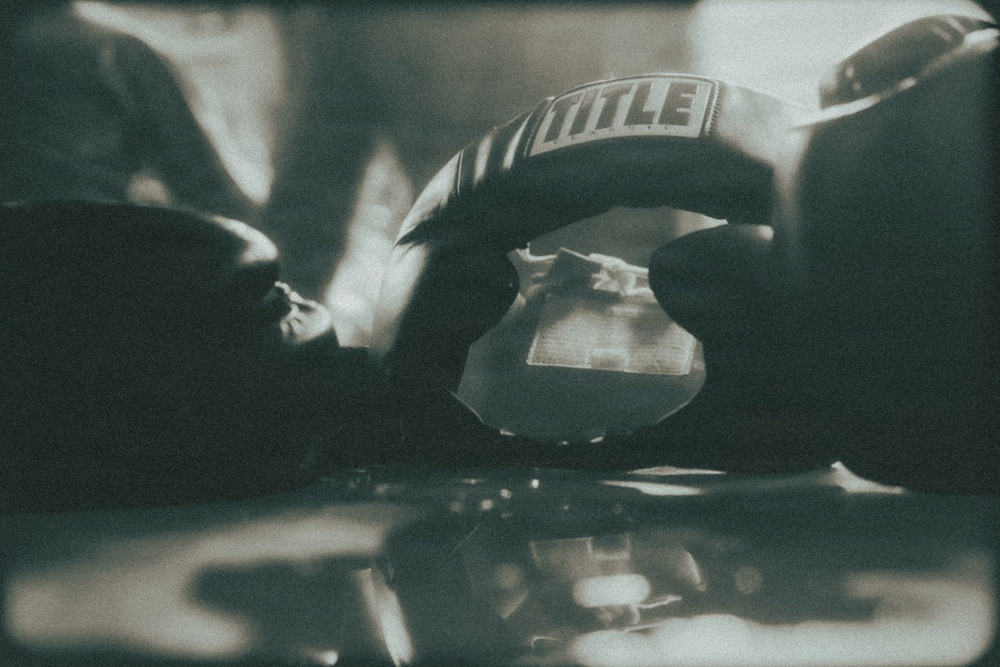 A close-up, moody photograph of boxing gloves bearing the 'TITLE' brand logo, resting on what appears to be a reflective surface. The image has a vintage, film-like aesthetic with a desaturated teal-green tone and shallow depth of field. The composition emphasizes the texture and weight of the gloves in an atmospheric gym setting.
