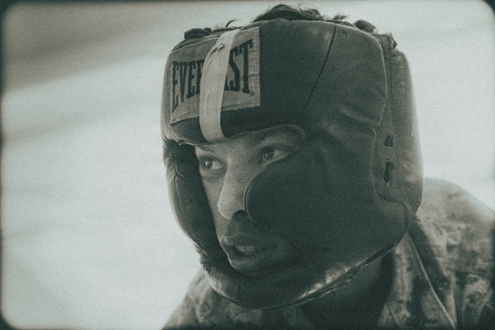 A close-up portrait of a person wearing an Everlast brand boxing headguard. The image has a vintage, grainy film aesthetic with a muted, desaturated tone. The subject appears focused and is wearing what looks like a camouflage or patterned garment.