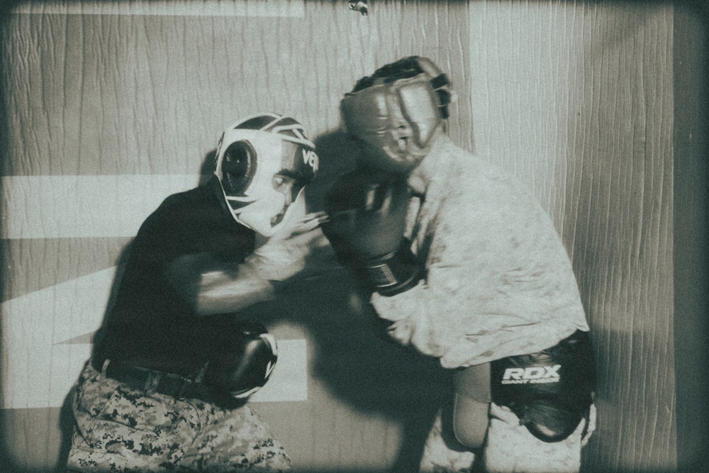 A monochromatic, film-grain stylized photograph showing two individuals sparring in a boxing or combat training session. Both participants are wearing protective headgear and boxing gloves, with one wearing camouflage military-style pants. The image has a vintage or aged aesthetic with visible light leaks and grain overlay.
