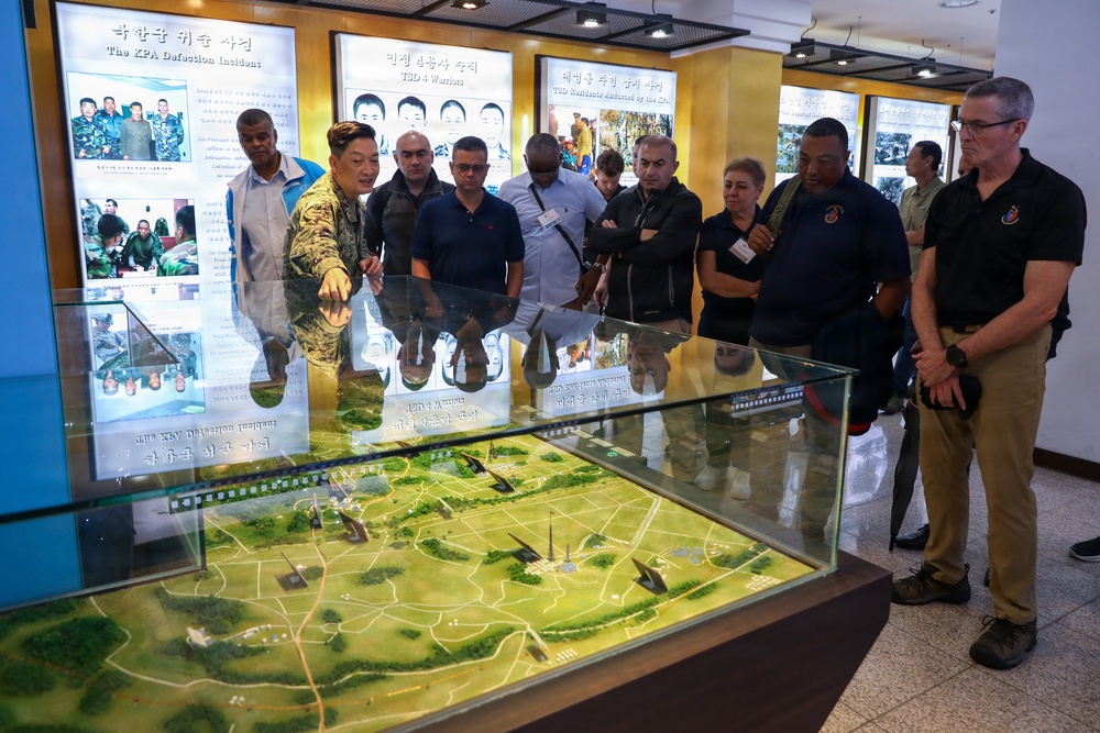 A military or government briefing is taking place at what appears to be a Korean DMZ or military museum exhibit. A person in military camouflage uniform is pointing at a detailed terrain diorama/scale model displayed in a glass case, while a diverse group of approximately 10-12 civilians and officials observe. The wall displays behind them contain bilingual (Korean and English) informational panels about incidents including 'The KPA Defection Incident' and 'USD Residents Abducted by the KPA'.