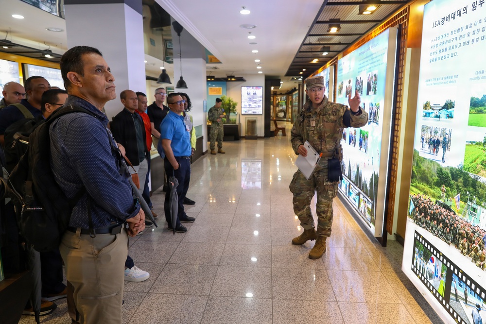 A U.S. military soldier in camouflage uniform is conducting a briefing or guided tour for a group of civilians and possibly other military personnel inside what appears to be a Korean military museum or visitor center. The soldier is gesturing toward a large display wall featuring Korean text and photographs. The setting includes Korean signage, suggesting this is likely at or near the Korean Demilitarized Zone (DMZ) or Joint Security Area (JSA).
