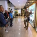 A U.S. military soldier in camouflage uniform is conducting a briefing or guided tour for a group of civilians and possibly other military personnel inside what appears to be a Korean military museum or visitor center. The soldier is gesturing toward a large display wall featuring Korean text and photographs. The setting includes Korean signage, suggesting this is likely at or near the Korean Demilitarized Zone (DMZ) or Joint Security Area (JSA).