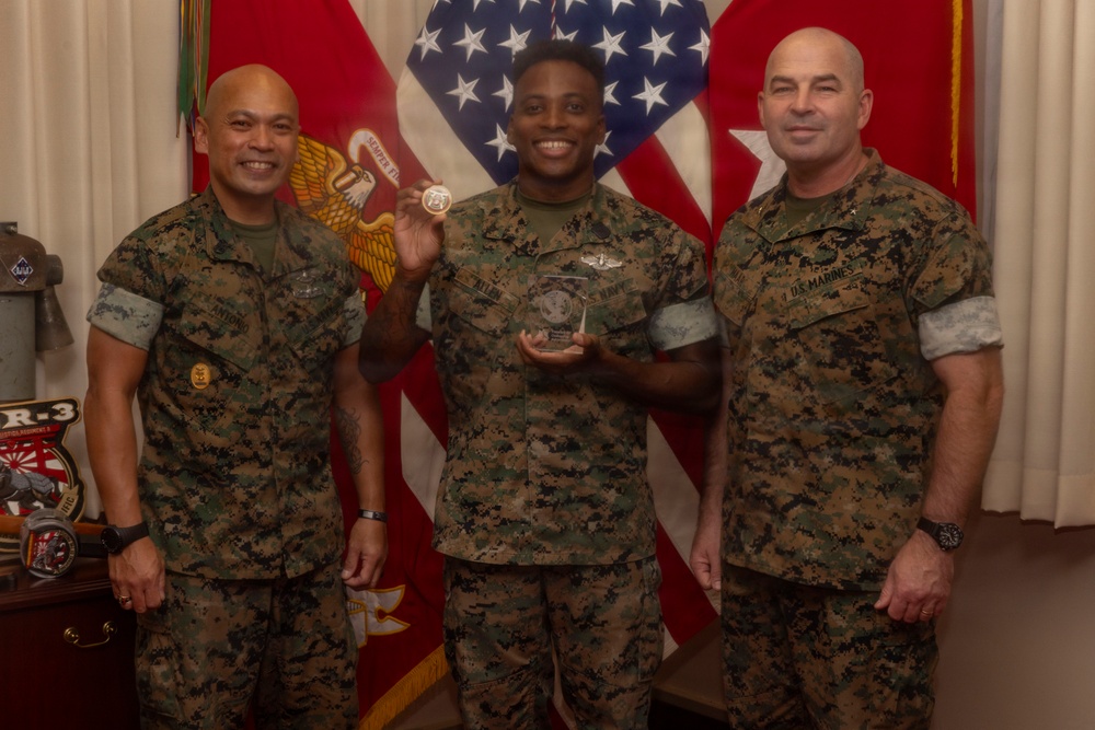 Three U.S. Marines in woodland MARPAT camouflage uniforms pose together in what appears to be an official recognition ceremony. The central figure is holding a challenge coin and a glass/crystal award trophy, suggesting he is being honored for an achievement. The background features the American flag and what appear to be U.S. Marine Corps unit flags or guidons.