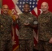 Three U.S. Marines in woodland MARPAT camouflage uniforms pose together in what appears to be an official recognition ceremony. The central figure is holding a challenge coin and a glass/crystal award trophy, suggesting he is being honored for an achievement. The background features the American flag and what appear to be U.S. Marine Corps unit flags or guidons.