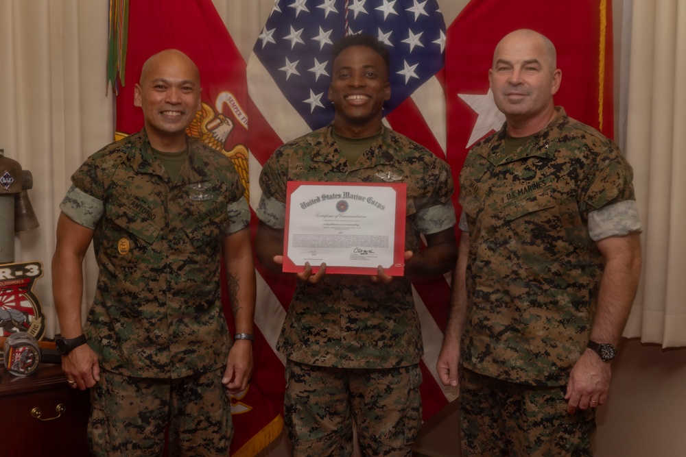 Three United States Marine Corps personnel are posing for a formal photograph. The central figure is holding a United States Marine Corps Certificate of Commendation with a red border. They are standing in front of American and Marine Corps flags in what appears to be an official office setting.